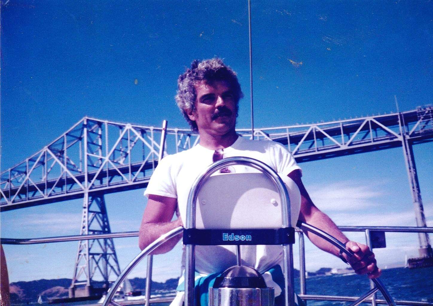 Photo of Jaime Jackson steering a sailing vessle on the San Francisco Bay, behind him visible the San Frnacisco Oakland Bay Bridge.